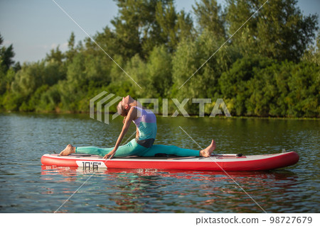 Young woman doing yoga on sup board with paddle. Meditative pose, side view - concept of harmony with the nature 98727679