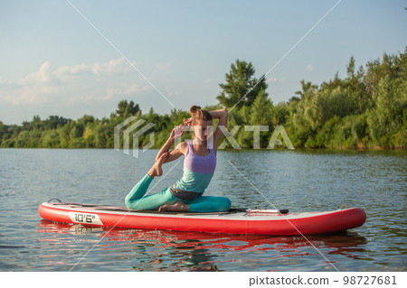 Young woman doing yoga on sup board with paddle. Meditative pose, side view - concept of harmony with the nature Young woman doing yoga on sup board with paddle. Meditative pose, side view - concept of harmony with the nature 98727681