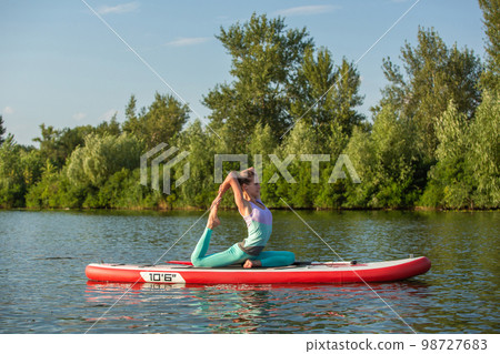 Young woman doing yoga on sup board with paddle. Meditative pose, side view - concept of harmony with the nature Young woman doing yoga on sup board with paddle. Meditative pose, side view - concept of harmony with the nature 98727683