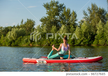 Young woman doing yoga on sup board with paddle. Meditative pose, side view - concept of harmony with the nature 98727685