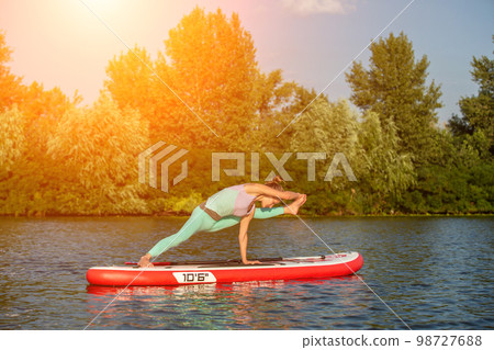 Young woman doing yoga on sup board with paddle. Yoga pose, side view - concept of harmony with the nature. Young woman doing yoga on sup board with paddle. Yoga pose, side view - concept of harmony with the nature. 98727688