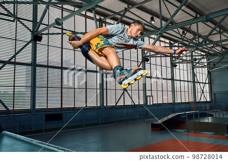 Rollerblader jump high from big air ramp performing trick. Indoors skate park equipment. 98728014