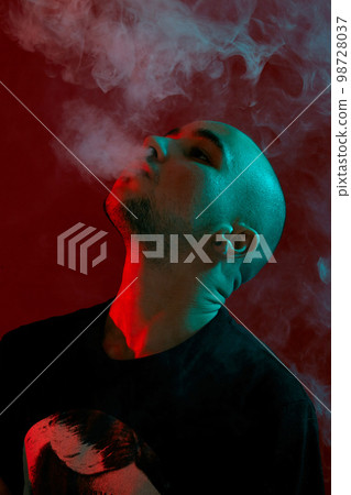 Close-up studio shot of a young bald guy vaping, blowing out a cloud of smoke on red background. Close-up studio shot of a young bald guy vaping, blowing out a cloud of smoke on red background. 98728037