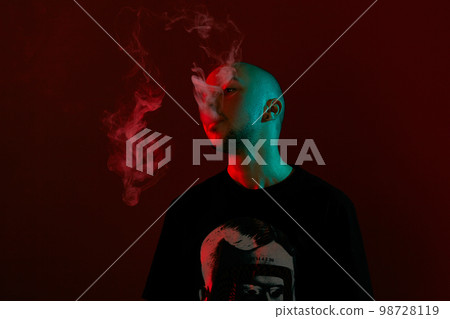Close-up studio shot of a young bald guy vaping, blowing out a cloud of smoke on red background. 98728119