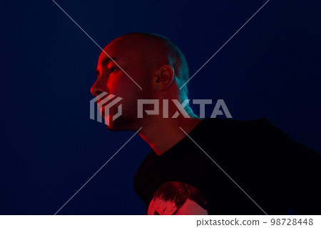 Studio shot of a young tattoed bald man posing against a blue background. 90s style. 98728448