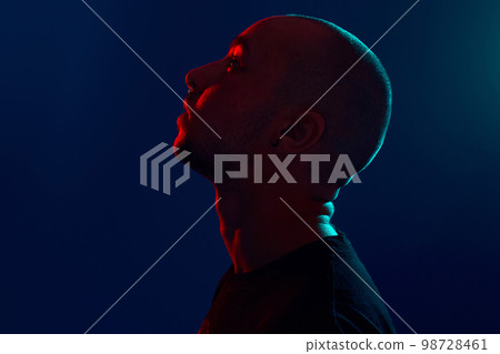 Studio shot of a young tattoed bald man posing against a blue background. 90s style. 98728461