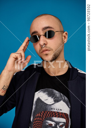 Studio shot of a young tattoed bald man posing against a blue background. 90s style. 98728502