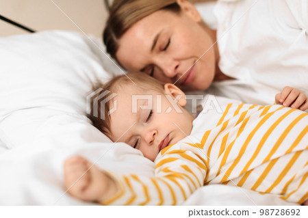Bed-sharing with baby concept. Adorable infant child girl sleeping with her mother on bed in bedroom during daytime Bed-sharing with baby concept. Adorable infant child girl sleeping with her mother on bed in bedroom during daytime 98728692