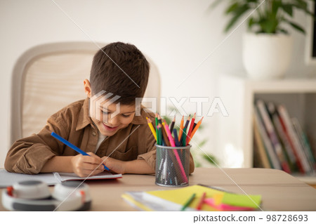 Busy cheerful cute small european boy sitting at table, draws picture in room, children garden or school interior Busy cheerful cute small european boy sitting at table, draws picture in room, children garden or school interior 98728693
