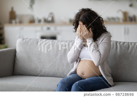 Portrait Of Young Pregnant Woman Crying While Sitting On Couch At Home 98728698