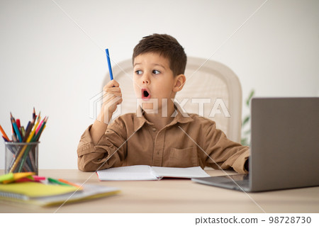 Happy surprised small european boy sit at table studying, drawing with laptop, looks at pencil with open mouth Happy surprised small european boy sit at table studying, drawing with laptop, looks at pencil with open mouth 98728730