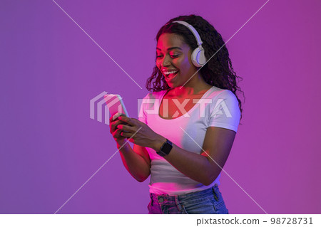 Excited Black Young Woman Wearing Wireless headphones Using Smartphone In Neon Light Excited Black Young Woman Wearing Wireless headphones Using Smartphone In Neon Light 98728731