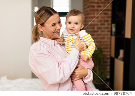 Motherhood concept. Happy mom with infant daughter on arms posing at home, kid looking at camera, free space 98728764