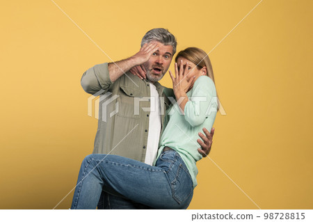 Portrait of scared middle aged couple covering mouth and eyes, woman jumping on man, looking at camera Portrait of scared middle aged couple covering mouth and eyes, woman jumping on man, looking at camera 98728815