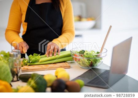 Cheerful millennial african american lady in apron making salad, cuts celery at table with laptop Cheerful millennial african american lady in apron making salad, cuts celery at table with laptop 98728859