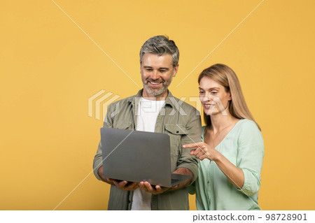 Excited middle aged caucasian couple using laptop together, woman looking and pointing at gadget, yellow background Excited middle aged caucasian couple using laptop together, woman looking and pointing at gadget, yellow background 98728901