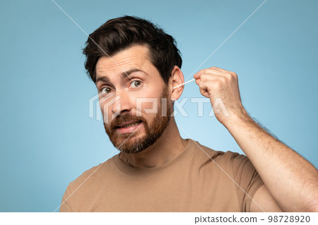 Funny middle aged bearded man cleaning ears with cotton swab, doing daily hygiene routine, blue studio background 98728920