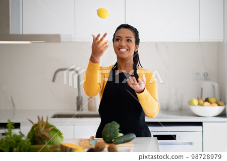 Cheerful millennial african american lady in apron juggles lemon, has fun at table with organic vegetables 98728979