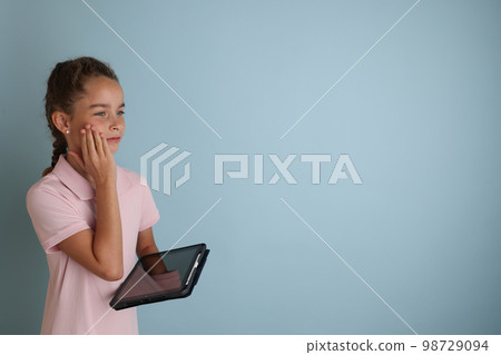 Little emotional teenage girl in pink shirt 11, 12 years old on an isolated blue background with a tablet in her hands.Children's studio portrait. Place the text to copy the place for the inscription Little emotional teenage girl in pink shirt 11, 12 years old on an isolated blue background with a tablet in her hands.Children's studio portrait. Place the text to copy the place for the inscription 98729094