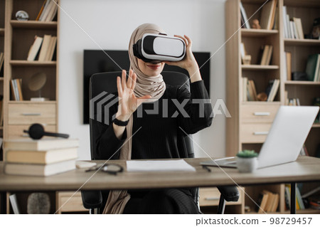 Muslim woman in headset touching virtual screen while sitting at desk with wireless laptop. 98729457