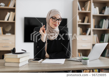 Smiling muslim woman in eyeglasses and hijab sitting at desk with books and notes. 98729895