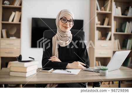 Smiling muslim woman in eyeglasses and hijab sitting at desk with books and notes. 98729896