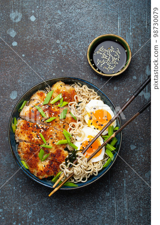 Asian noodles ramen soup with deep fried panko chicken fillet and boiled eggs in ceramic bowl with chop sticks, soy sauce on stone rustic background top view Asian noodles ramen soup with deep fried panko chicken fillet and boiled eggs in ceramic bowl with chop sticks, soy sauce on stone rustic background top view 98730079