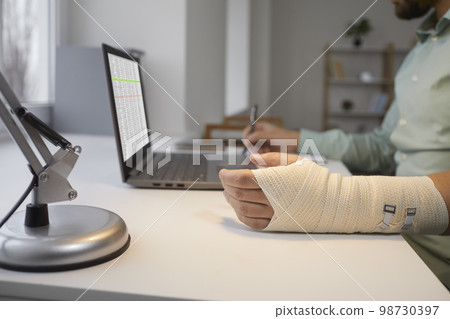 Bearded man with bandaged broken arm is working at laptop, sitting at his desk and making notes in notebook for reporting. 98730397