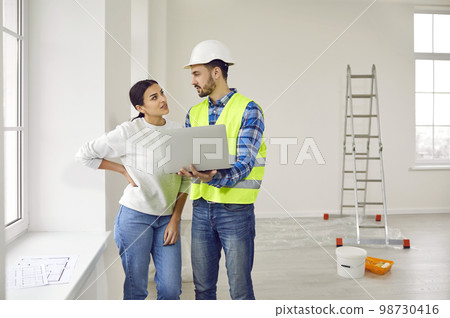 Attractive young woman is talking to construction foreman in white helmet, who demonstrates sequence of repairs in living room on laptop. 98730416