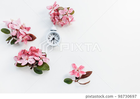 Small white clock with bell on white with fresh spring daisy flowers background. Spring forward, daylight saving time in March concept 98730697