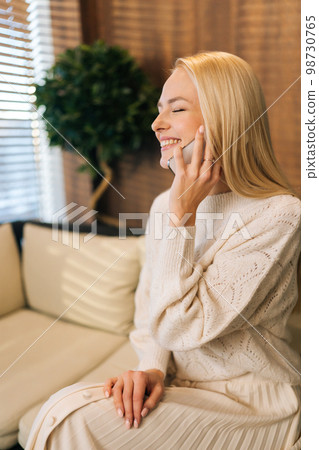 Vertical side view shot of joyful young woman talking on mobile phone sitting alone on sofa by window in cafe, closed eyes. Happy blonde female conversation with smartphone while rest during breakfast 98730765