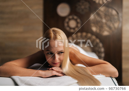 Close-up portrait of pretty young woman lying on massage table in wellness center for massage therapy. Beautiful blonde female lying down on therapeutic table at spa salon awaiting beauty treatment. 98730766
