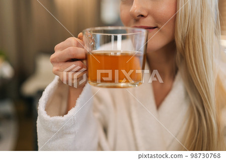 Close-up cropped shot of unrecognizable blonde woman wearing white bathrobe drinking hot healthy herbal tea enjoying spa procedures in spa salon. Concept of rest with spa treatment. Close-up cropped shot of unrecognizable blonde woman wearing white bathrobe drinking hot healthy herbal tea enjoying spa procedures in spa salon. Concept of rest with spa treatment. 98730768