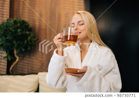 Happy excited young woman in white bathrobe enjoying drinking hot healthy tea with closed eyes after spa procedures. Calm blonde female resting with herbal drink sitting in sofa at spa salon. 98730771
