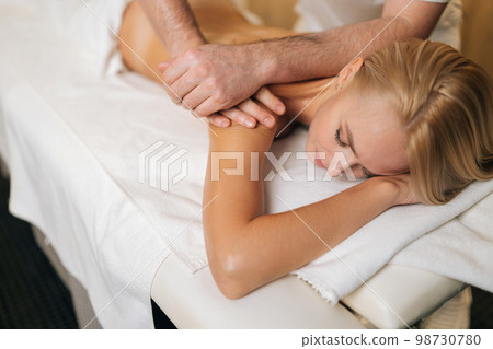 Cropped shot of unrecognizable professional male masseur therapist doing wellness beauty treatment to attractive blonde female lying on treatment table in spa salon. Concept of healthy lifestyle. Cropped shot of unrecognizable professional male masseur therapist doing wellness beauty treatment to attractive blonde female lying on treatment table in spa salon. Concept of healthy lifestyle. 98730780