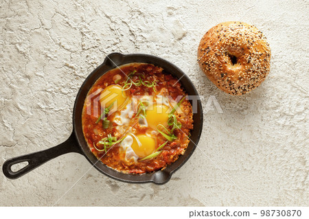 Shakshuka in cast iron pan with freshly baked bagel on white textured background. Fried eggs with tomato sauce. Shakshuka a traditional meal of the Jewish cuisine 98730870