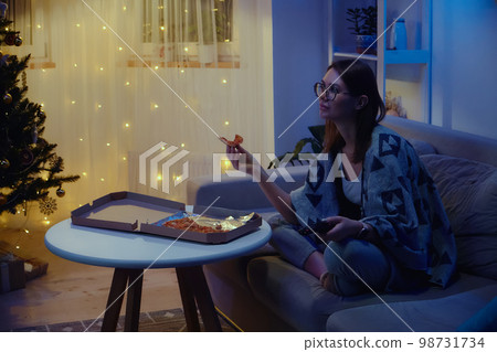 Happy woman eating pizza alone while watching tv at christmas on christmas tree background 98731734