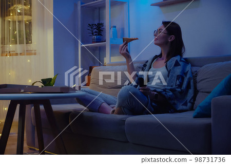 Happy woman eating pizza at night watching tv while sitting on sofa at home 98731736