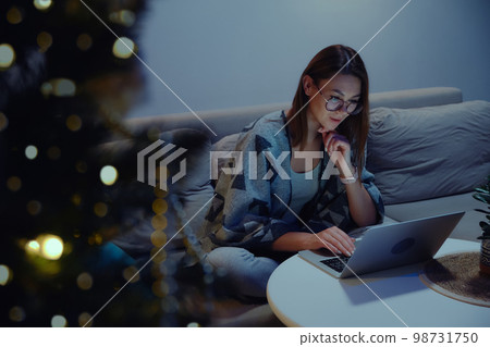Young smiling woman using laptop at night or evening, video call sitting on sofa at home against background of christmas tree and lights Young smiling woman using laptop at night or evening, video call sitting on sofa at home against background of christmas tree and lights 98731750