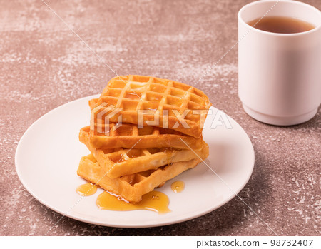 Delicious Belgian waffles with honey served on white plate and brown background, cup of black tea, rustic concept. Delicious Belgian waffles with honey served on white plate and brown background, cup of black tea, rustic concept. 98732407