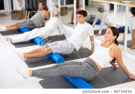 Young positive female performing set of exercises with pilates roller during group class in fitness studio. 98734830