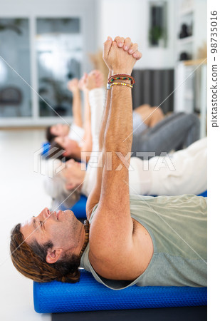 Middle-aged man practicing pilates with roller on gray mat in gym room 98735016