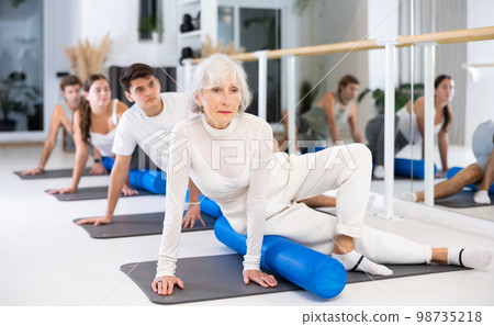 Active senior woman performs exercises with a pilates roller while lying on a mat in the hall of a modern fitness studio. Active senior woman performs exercises with a pilates roller while lying on a mat in the hall of a modern fitness studio. 98735218