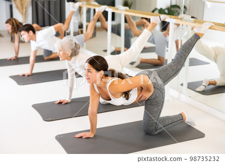 Positive young female performing set of exercises on a pilates mat during group class in fitness studio. 98735232