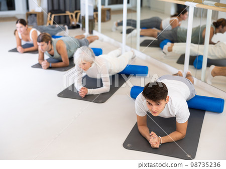 Positive athletic male performing set of exercises with pilates roller during group class in fitness studio. 98735236