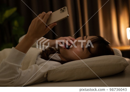 A woman operating a smartphone while yawning in the bed/bedroom 98736043