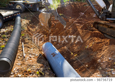 Worker operating tractor lift sewage pipes in ground is preparing to install underground concrete pipes 98736719
