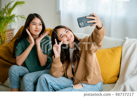 Two asian young women happy smiling and taking selfie on couch in living room at home 98737217