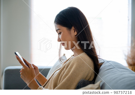 Happy young asian woman relax on comfortable couch at home texting messaging on smartphone 98737218