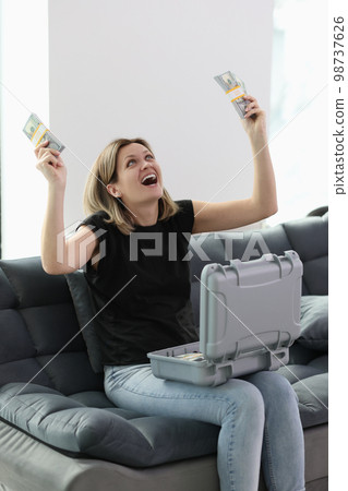 Happy woman holding stacks of dollar bills in hand while sitting on sofa with suitcase full of money Happy woman holding stacks of dollar bills in hand while sitting on sofa with suitcase full of money 98737626